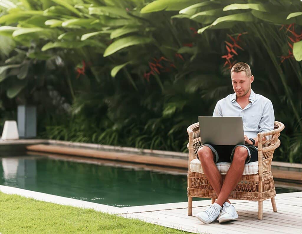 Man sitting poolside in wicker chair working on laptop